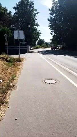 The video shows a pavement beside a road with trees on either side. There is a manhole cover on the pavement. There are traffic signs on a pole on the left side of the pavement. A car is visible in the distance on the road. The sky is blue and there are some clouds. The sun is shining. The pavement is made of concrete and the road is made of asphalt. There is some dry grass on the left side of the pavement. The video is taken from a first-person perspective.