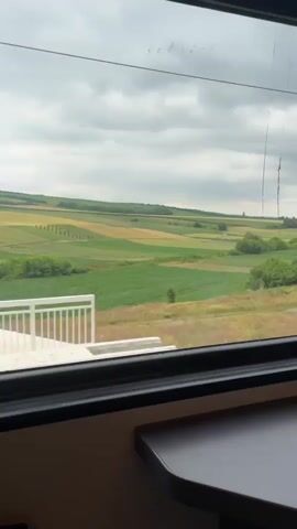 A video of a view from a train window shows a vast landscape of green and yellow fields under a cloudy sky. The train appears to be moving, as the landscape outside shifts from left to right. In the foreground, there are bushes and trees, with a dirt path visible between them. In the distance, more fields and scattered trees can be seen. Inside the train, a dark brown table is visible in the lower right corner, with the window frame visible at the bottom and sides of the shot. The lighting is natural, suggesting daytime.