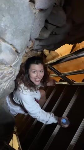 A woman smiles and takes a sip from a water bottle while climbing up a dark set of wooden stairs, enclosed by rock and metal railings. She is wearing a white sweatshirt, a white skirt with black stripes, and a red heart-shaped purse slung across her shoulder. The rough texture of the rock walls creates a cave-like atmosphere.