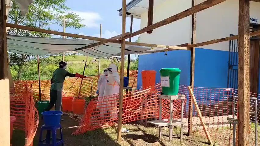 This media file captures an outdoor decontamination station under a bright, sunny sky.

**Subjects:** Three individuals are prominently featured. One person, dressed in green scrubs or workwear, a face shield, and dark gloves, operates a handheld sprayer. They are actively spraying two other individuals who are fully encapsulated in white personal protective equipment (PPE) suits, complete with hoods, masks, and gloves. A fourth person, also in green scrubs, is partially visible on the far left of the frame.

**Objects:** The decontamination area is constructed with a makeshift wooden frame, partially covered by a white tarp providing shade. The perimeter is demarcated by orange plastic safety netting. Various plastic containers are scattered around the ground: several orange buckets, a blue bucket, a green bucket, and a black one. A green bin, labeled "LAVAGE MAINS" (Hand Washing), rests on a wooden stand. Another orange bin is visible further back. In the background stands a building with a blue lower wall and a white upper wall, featuring a door and a window.

**Scene & Setting:** The scene takes place outdoors on a dirt or grassy patch, possibly adjacent to an institutional building. The improvised nature of the decontamination station suggests a temporary setup in response to a public health or environmental safety need.

**Activity:** The primary activity is the disinfection or decontamination of individuals wearing protective suits. The person in green is spraying the two individuals in white PPE, likely to clean or neutralize any contaminants on their suits after they have exited a potentially hazardous zone.

**Notable Details:**
*   **Time of day:** Daytime, with bright sunshine casting clear shadows, indicating good weather.
*   **Weather:** Clear and sunny.
*   **Location Cues:** The makeshift construction and general appearance suggest a location in a developing region. Given the context of Tchere, Democratic Republic of the Congo, this setup likely serves as an Ebola or other infectious disease response point, or a similar emergency health measure.
*   **Visible Text:** The green bin clearly displays "LAVAGE MAINS" (Hand Washing). Faint text is also visible on the white upper part of the building in the background, possibly "MAGASIN" (Store/Warehouse).
*   **Emotions:** The individuals' faces are largely obscured by PPE, but the activity appears to be conducted in a focused and procedural manner.