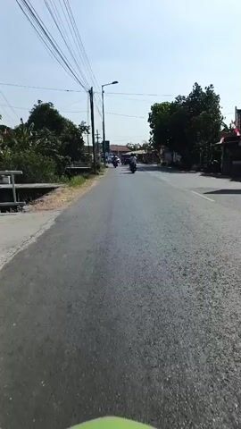 The video shows a first-person perspective of someone riding a scooter down a paved road. The sky is clear and blue, indicating it is daytime. On the left side of the road, there is a ditch with some vegetation and trees. On the right side of the road, there are more trees and some buildings, including a house with a red roof. There are several other vehicles on the road, including motorcycles and cars. The scooter rider is moving forward, following the road. The scene appears to be in a rural or suburban area.