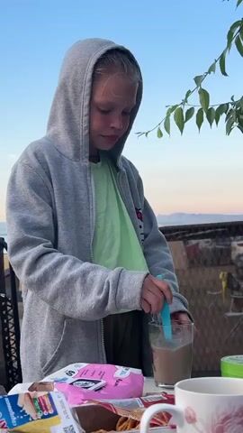 A young girl wearing a grey hooded sweatshirt over a green shirt and dark pants stands outside at a table, stirring a glass of brown liquid with a blue stirrer. Her hair is blonde and pulled back. In front of her on the table are several food items, including a pink package, a box of crackers, and a white mug with a floral design. Behind the girl, there is a blurred background of trees, a fence, and a body of water. The sky is a pale blue with some pink hues, suggesting it is either sunrise or sunset.