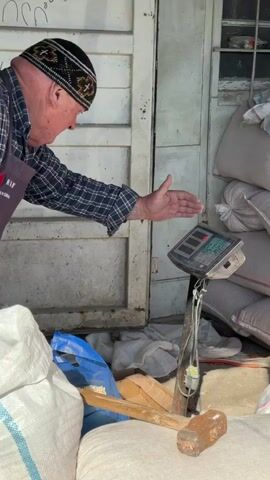 An elderly man is seen in what appears to be a market or storage area. He is wearing a checkered shirt, an apron with the text "RAW, trading in Tbilisi," and a traditional black cap. He is bending over and interacting with a digital scale. He places a green scoop on the scale and then appears to be taking something out of a blue plastic bag. There are several white sacks filled with goods behind and around him, along with a wooden tool and a brick on top of one of the sacks. The background includes a white door or wall with some writing in Georgian. The lighting is natural and the setting appears to be somewhat cluttered.