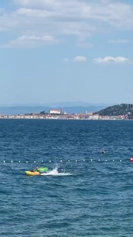 This is a video clip of a beach scene on a sunny day. The camera is positioned low, looking out over a blue ocean. In the foreground, there are a few people in the water. One person is on a yellow and green inflatable float, kicking their legs and creating a splash. Another person is further out in the water, swimming. A red buoy is visible in the distance.

In the background, a coastal city with red-tiled roofs and a prominent church tower is visible. The city is nestled on a hillside that slopes down to the sea. The sky is a clear blue with a few white, fluffy clouds. The overall impression is one of a pleasant summer day at the beach.

The video appears to have been filmed during daylight hours, given the bright sunlight. The presence of people swimming and enjoying water activities suggests a warm weather climate. The distinctive architecture of the city in the background, combined with the red-tiled roofs, is characteristic of many coastal towns in the Mediterranean region. The specific mention of Crveni Vrh, Croatia, provides geographical context, indicating the location is likely on the Istrian peninsula. There is no visible text in the video. The mood is relaxed and recreational.