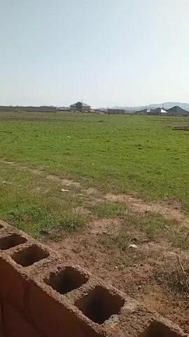 The video shows an outdoor scene with a grassy field and a dirt path. In the foreground, there is a pile of debris including plastic bottles and other trash. The path is lined with more trash. In the middle ground, there is a fence made of bricks and a building behind it. The background features a vast green field with some scattered trees and buildings in the distance, and hills or mountains under a bright, clear sky. The video appears to be a panning shot, moving from left to right.