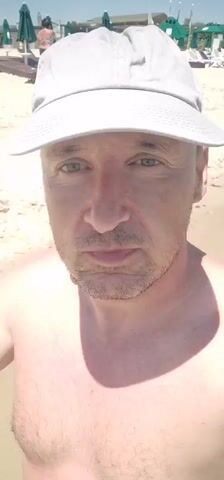A man wearing a white baseball cap and no shirt stands on a beach, holding a camera, and filming himself. The man's face is in focus. In the background, the beach is mostly empty with a few lounge chairs and umbrellas visible. The ocean is in the distance. There are a few other people in the background, but they are out of focus and too far away to be clearly identified.