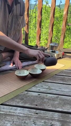 A person sits cross-legged on a wooden deck, pouring tea from a dark teapot into three small, light-colored bowls arranged in a line. The bowls are on a woven mat. In the background, a portable stove with a kettle sits on the deck. Green trees and a mountain are visible beyond the wooden railing. The person wears a plaid shirt and dark pants.