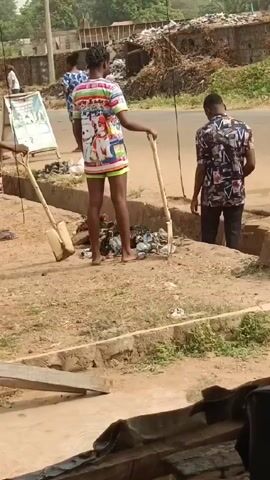 The video shows three individuals standing near a ditch filled with trash on the side of a road in what appears to be a developing country. One person, dressed in a colorful top and shorts, stands holding a shovel. Another person is in the ditch, and a third person is walking towards the ditch carrying a large metal platter. A pile of trash is visible in the background. The overall environment appears to be a busy roadside with signs and other people present.