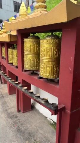 A row of golden Buddhist prayer wheels are shown. They are cylindrical and have an inscription in an unknown language running around them. They are mounted on a red wooden frame, under a yellow roof. In front of the prayer wheels, there is a red shelf with a row of small brass oil lamps. The ground in front of the shelf is paved with rough stones and dirt. Behind the prayer wheels, there is green foliage.