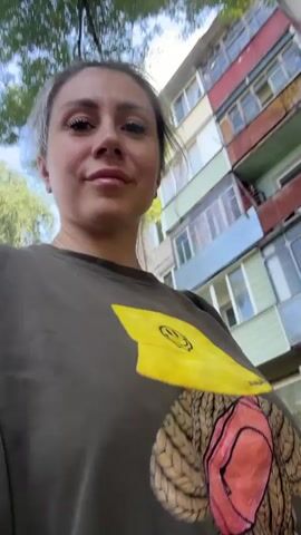 The video shows a close-up, slightly shaky, first-person perspective of a woman with short, dyed blonde hair and light makeup, looking up and around. She is wearing a dark green sweatshirt with a large yellow graphic on it, featuring a smiley face and what appears to be abstract artwork. In the background, a weathered, multi-story apartment building with balconies and various colored sections is visible, along with green trees. The lighting suggests it is daytime. The woman briefly turns her head and looks towards the camera with a neutral expression before the video ends.