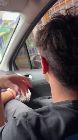 The video offers a rear perspective of a young man seated in the front passenger seat of a car. He wears a black t-shirt and grey pants, with his dark, medium-length hair visible. His left leg is propped up, displaying a white and grey sneaker with a subtle red accent near the ankle. The man is looking out the right-side window, which reflects portions of the car's interior, including the dashboard and a rearview mirror.

Outside the car, an urban or suburban landscape unfolds under bright daylight. Colorful building facades, possibly residential or commercial structures, are visible in the background, suggesting a lively environment. The car appears to be either stationary or moving at a very slow pace. The man's left hand is partially seen, holding what might be a small object or a phone, resting near his propped leg. The scene takes place in Ramos Mejía, Argentina. No specific emotions are discernible from this viewpoint, and there is no visible text within the frame. Muffled speech, possibly in Spanish, is audible in the background.