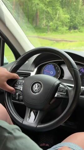 The video shows the interior of a car, specifically the steering wheel and dashboard, from the perspective of the driver. A person's hands are visible, one resting on the steering wheel and the other pressing buttons on the left side of the wheel. The steering wheel is black leather with a chrome Volkswagen logo in the center. The dashboard displays a circular speedometer with a needle indicating a speed, and a digital display showing "166882" and "30". The background visible through the windshield is a blur of green trees and foliage, suggesting an outdoor, possibly rural or wooded setting. The lighting indicates daytime, and the weather appears to be overcast or cloudy, as there are no harsh shadows. The visible text on the dashboard includes "AIRBAG" on the steering wheel, and numbers and text on the digital display, some of which are in Cyrillic script ("Дистанция" and "Среднее"). The overall mood is calm and focused on the act of driving or interacting with the car's controls. The location context indicates Cheposh, Russia.