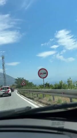 The video shows a point-of-view shot from inside a car driving on a road. The road has a white line marking the edge and a metal guardrail. There is a red triangular warning sign with a double curve symbol. Ahead, a white car with red accents is visible. The landscape includes green trees and bushes on the side of the road, and in the distance, a body of water, possibly the sea, and hills. The sky is blue with scattered white clouds.