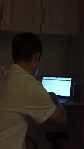 A person wearing a white t-shirt is sitting at a desk, facing a laptop. They are typing on the keyboard. The laptop screen is illuminated. To the right of the laptop, there is a dark bottle. Above the desk, there are light-colored cabinets with gold handles. The lighting in the room is dim.