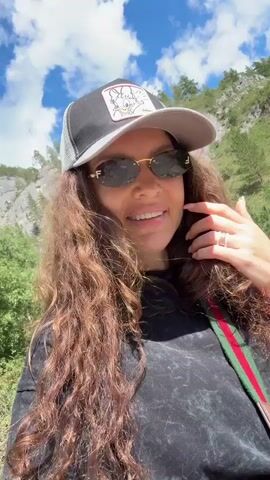 A young woman with long curly brown hair, wearing a black t-shirt, a gray and black baseball cap, and sunglasses smiles and poses for the camera. She has on jewelry including rings and a watch. A crossbody bag with a green and red stripe is visible. The background features a mountainous landscape with green trees and a blue sky with white clouds.