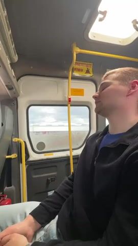 A man sits on a bus with his eyes closed, apparently asleep. He is wearing a black jacket and blue jeans. The bus appears to be traveling through a rural area with several farm buildings visible through the window. The sky is overcast.  A yellow handrail is positioned near the window.