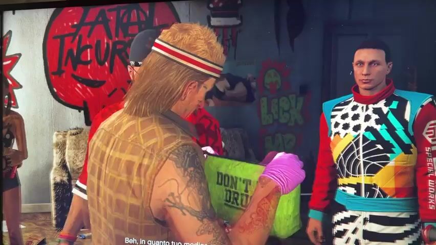 The video shows three men in what appears to be an indoor setting with graffiti on the walls. In the foreground, a man on the left with a red shirt, black bandana, glasses, and a beard is looking down. In the center, a man with blonde hair, a headband, and a plaid vest is wearing pink gloves and holding a green bag. He is looking into the bag. To the right, a man in a colorful jacket with black and white polka dots is watching. The man in the center appears to be interacting with the contents of the green bag. The overall scene suggests a clandestine transaction or exchange.