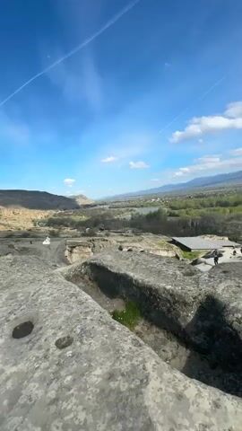 The video presents a wide shot of a rocky landscape with cut stone features in the foreground. The sky is bright blue with a few streaky clouds and jet contrails. In the distance, a river flows through a green valley, with hills and mountains visible on the horizon. Several small figures of people can be seen walking in the middle distance. The rock in the foreground contains a few cylindrical holes and a rectangular excavation where some green plants are growing. The shadow of a person holding a mobile phone camera is visible in the bottom left corner of the frame.
