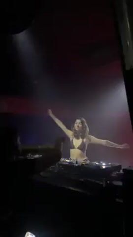 Here is a description of the video:

A dimly lit DJ booth is shown in a nightclub. A light shines on the female DJ who stands behind her equipment, wearing a black bikini top. She raises her arms while the music plays and she smiles. Another person stands at the corner of the booth, facing the DJ, with their arm raised. The room has dark walls, and there are colored lights.