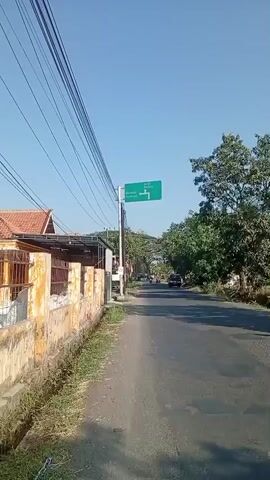 A green directional sign with white text and arrows is mounted on a metal pole. The text on the sign reads "Wonorejo Purwosari" with an arrow pointing left, and "Bangil Rembang" with a T-junction symbol indicating a right turn. The sign is positioned alongside a paved road. To the left of the road, there is a low wall and some vegetation. Several utility wires are visible overhead. In the background, there are trees, a few vehicles, and buildings. The sky is clear and blue.