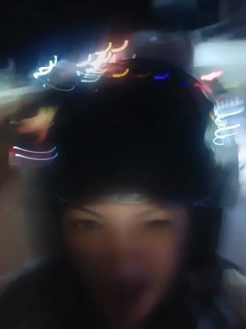 A first-person perspective video shows a person wearing a black helmet and a light-colored jacket or shirt, possibly riding a motorcycle at night. The background is blurred due to motion and low light, but appears to be a street scene with other vehicles and illuminated signs. There are several bright headlights from oncoming vehicles, creating a streaking effect. The person in the video appears to be looking at the camera and the scene is dynamic, indicating movement. Orange traffic cones or barriers are visible on the side of the road. A billboard is also visible in the background.