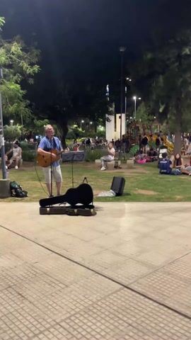 At night, a man is playing the guitar and singing in a park. He is wearing a blue short-sleeved shirt and beige shorts and is standing on a stone walkway.  In front of him is an open guitar case, a speaker, and a music stand. Behind him is a large grassy area, illuminated by streetlights, where people are sitting on the grass and walking around. A small playground is also visible in the background.