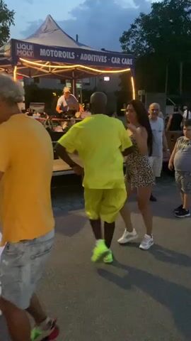 A group of people are gathered outdoors at dusk, possibly for an event. In the foreground, a man in a yellow t-shirt and shorts with bright green shoes dances with a woman in a leopard-print dress. To the left, another person wearing a yellow shirt and denim shorts is visible from behind, also appearing to be dancing. Behind the dancing couple, a stage is set up with a DJ playing music and a tent overhead that displays "LIQUI MOLY MOTOROILS ADDITIVES CAR CARE". Several other people are standing around watching or participating in the event. The ground is paved, and string lights are visible around the stage. The sky is dark blue, suggesting it is evening.
