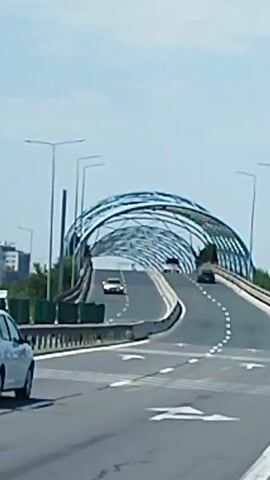 A vertical video shows a white sedan driving on a highway under an arched bridge. The bridge has a modern, metallic design with a curved, lattice-like structure. The highway has multiple lanes with white lane markings and directional arrows painted on the asphalt. Other cars are visible on the highway, some in front of and behind the white sedan. The sky is clear and blue, suggesting a daytime scene. Traffic signs are visible on the right side of the road, including a pedestrian crossing sign and a blue circular sign with white symbols. The environment appears to be an urban or suburban area, with some buildings and greenery visible in the background.