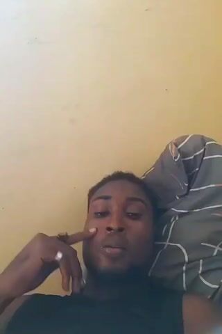 The video shows a man lying on a bed, looking at the camera. He is wearing a black sleeveless shirt and has short black hair. He raises his arm and puts his hand behind his head. The bed has gray and white striped sheets. The wall behind him is yellow.