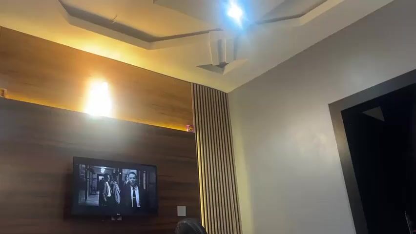 The scene is a brightly lit room. The main focal point is a large, flat-screen TV mounted on a wooden panelled wall. The TV is showing a black-and-white film featuring two men in suits. Adjacent to the wooden panel is a section of vertical wooden slats. The wall to the right of this is a plain, light grey colour. A black-framed doorway is positioned to the right. The ceiling is white with a recessed, geometric design and a bright light fixture in the centre. The room appears to be a living space. The image is static.
