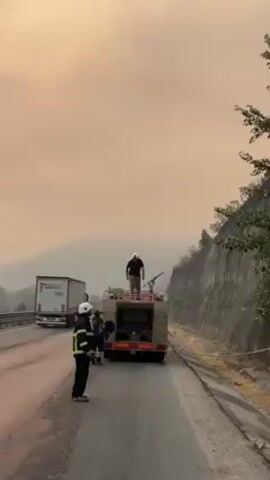 The video shows a fire engine on the side of a road, with a firefighter standing on top of it, tending to the equipment. Two other firefighters are standing beside the vehicle. A truck drives past on the road, and there is a concrete wall and trees on the other side of the road. The sky is obscured by smoke or fog, giving it an orange hue.