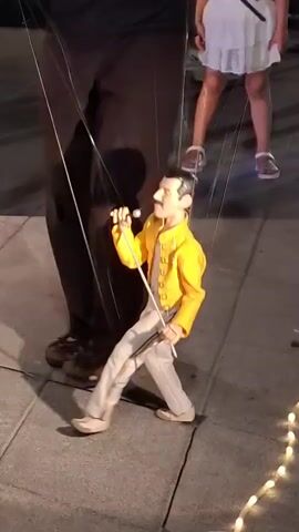The video displays a puppet resembling Freddie Mercury, dressed in a yellow jacket and white pants with rainbow stripes. The puppet is being manipulated by strings, controlled by unseen puppeteers. In the background, a person's legs and feet are visible, and to the left, a small toy piano and a gold curtain are present. The puppet is performing, holding a microphone in its right hand and moving its arms as if singing or playing an instrument. The scene appears to be outdoors on a paved surface.