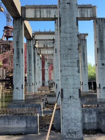 This is a video of an abandoned industrial site, likely a power plant, on a sunny day. The camera pans across a row of imposing concrete pillars, revealing the immense scale of the structure. In the background, a large, rusty building dominates the scene, its metal panels weathered and stained. A collapsed bridge or gantry lies partially submerged in the murky water below, adding to the sense of decay and abandonment. Trees and greenery are beginning to reclaim the site, with vegetation growing on the rooftop of the rusty building and along the water's edge. The sky is a clear blue, suggesting good weather. The overall atmosphere is one of solemn grandeur and the poignant beauty of nature slowly taking over human-made structures. There is no visible text in the video. The location is identified as Kyiv, Ukraine.