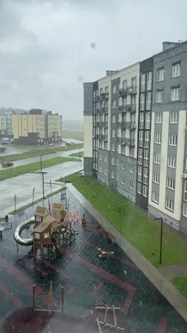 This is a vertical video of a playground and an apartment building on a rainy day. The playground is located in front of the apartment building and has a slide, swings, and other play structures. The apartment building is a modern, multi-story building with balconies and many windows. The sky is cloudy, and there are visible raindrops falling. The ground is wet, indicating that it has been raining for some time. The overall scene is somewhat gloomy due to the weather.