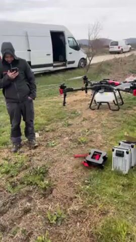 A man stands beside a large agricultural drone, and someone is looking at a handheld device, probably controlling the drone. The drone is equipped with a tank, presumably for spraying crops. Nearby, there are batteries and some other equipment. The man is wearing a black jacket with "EA7 EMPORIO ARMANI" on the chest and a "BOSS" shirt underneath.  A survey pole is set up in a field. The sky is overcast and there are two white vans parked in the background. The video is likely about agricultural spraying using drone technology.
