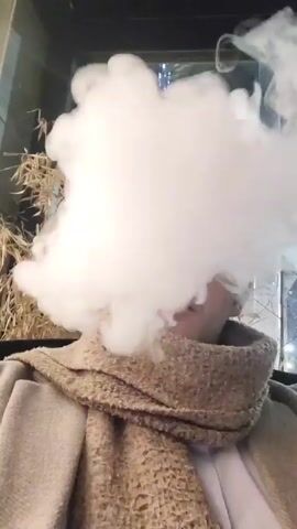 The video shows a person surrounded by a large cloud of white vapor. The person is wearing a beige coat, a scarf, and a white shirt. The background appears to be an outdoor seating area with plants and lights visible. The vapor obscures the person's face.