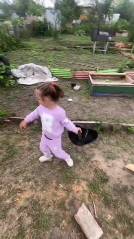 A young girl in a light purple tracksuit is in a yard, holding a large blue jug in her left hand. She turns and begins to swing the jug in a circular motion. The jug is then replaced with a black shovel. The girl appears to be interacting with the shovel as if it were a toy. The yard is dirt and grass with some scattered debris, including a white tarp, green corrugated metal sheets, and a wooden sandbox. In the background, there is a grill, trees, and a house.