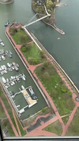 The video shows an aerial view of a river and coastline area, likely a city or town. The camera is positioned inside a building, peering through a window. 
The foreground features a marina with multiple docked boats. A landscaped peninsula is visible, featuring green spaces, walking paths lined with red paving, and a white bridge connecting to a small island. The island includes a beach area. 
In the background, a city skyline is visible under an overcast sky, including industrial buildings and other cityscapes. The river is wide and appears to flow into the distance. A ship is moving on the river.
There are indistinct voices and sounds in the background, which suggests there are people nearby.