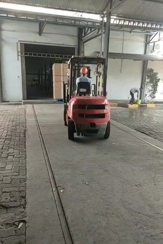 A red forklift with a person wearing a blue shirt and an orange helmet is driving towards the left on a concrete surface. The forklift is carrying boxes. To the right, another person wearing a grey shirt and blue pants is standing and holding a green bucket. The environment appears to be an outdoor loading dock area with a metal roof structure overhead.