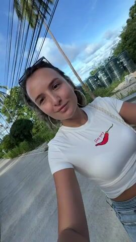 A woman with brown hair and hoop earrings wears a white t-shirt with a chili pepper graphic and the words "Stay Spicy" printed on it. She is walking on a paved road with a yellow line on the left side. There are power lines above and to the left, with lush green trees and vegetation on the right side of the road, as well as a white metal fence. The sky is blue with scattered clouds. The woman is looking at the camera and making a kissing motion.