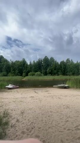 A person is swimming in a body of water near a sandy beach. There are two boats on the water. One boat is white with red accents and is closer to the shore on the left side of the frame. The second boat is green and white and is further out in the water on the right side of the frame. The beach is sandy with some sparse vegetation. Behind the beach is a line of dense green trees. The sky is cloudy.