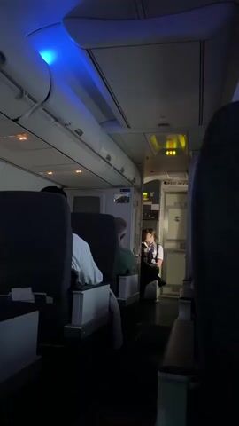 The video shows the interior of an airplane cabin with dim lighting. There are several rows of seats visible, some occupied by passengers. A flight attendant is seated in the galley area at the rear of the cabin. The cabin lighting is predominantly blue and yellow.