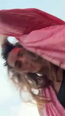 The video shows a first-person perspective of someone at an outdoor event, possibly a festival. The person is wearing a pink shawl or garment that initially covers the camera. The camera is then moved, revealing a crowd of people and a person in the background holding a large, colorful, round balloon or kite. The people appear to be moving or dancing, and there's music playing in the background. The person holding the camera makes gestures towards the camera with their hand. The environment includes trees and a slightly overcast sky.