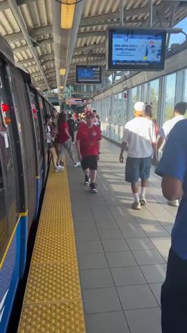 The video is a first-person perspective video of a train station platform. A train is stopped at the platform on the left side of the frame. Several people are standing on the platform, some of whom appear to be boarding or disembarking the train. The platform has a yellow tactile warning strip along the edge. Overhead, there are lights, signs, and screens displaying information. The environment appears to be an elevated or covered train station with a view of the outdoors through large windows on the right. A man in a blue shirt and a baseball cap is walking away from the camera towards the right side of the frame. A person in a red shirt with a Canadian flag logo is walking towards the camera. Other individuals are scattered along the platform. The lighting suggests it is daytime.