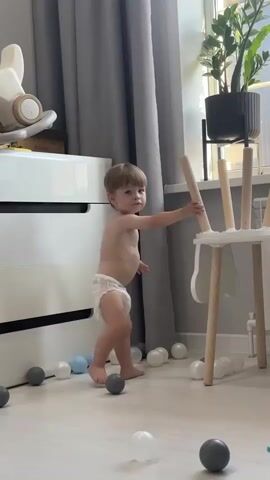 A baby wearing a diaper stands in a room with light-colored flooring and grey curtains in the background. The baby is facing forward and appears to be looking at the camera. Scattered on the floor around the baby are several small, colorful balls, mostly grey and white, with one blue ball visible. To the left, a white dresser with black drawers is partially visible, and on top of it is a light-colored toy with wheels. To the right, a white chair with wooden legs stands near a window, and a potted plant with green leaves is placed on a shelf or stand near the window. The baby is in the center of the frame, with its arms slightly outstretched and legs apart, in a stance that suggests it is about to move or has just stopped. The lighting in the room appears to be natural, coming from the window.