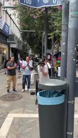 The video shows a man busking in the street. He stands on a paved sidewalk next to a black garbage receptacle and is rapping or singing into a microphone. He’s wearing dark trousers, a light-colored shirt, and a dark baseball cap, and sunglasses. To the left of the busker, two men clap their hands and appear to be enjoying the show. One man is wearing a black T-shirt and khaki cargo pants, while the other wears a white patterned shirt and jeans. Another woman stands behind the clapping men, smiling. Behind them, the street is busy with pedestrian traffic. Buildings with storefronts line either side of the street, and trees overhang the street, with flowers visible. A blue street sign is affixed to a post at the right of the screen, reading “100”.
