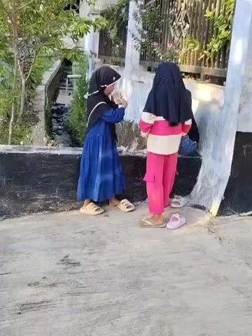 Two young girls wearing hijabs stand near a wall and a fence outdoors. The girl on the left wears a blue dress and tan sandals. The girl on the right wears pink pants, a striped shirt, and pink sandals. They appear to be interacting with each other, possibly playing or talking. The background shows greenery, a building, and a ditch or water channel. The ground is paved.