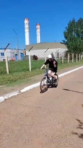 A young person is seen riding a blue and silver mountain bike on a paved road. They are wearing a black shirt, black shorts, a white baseball cap, and a black backpack. The person makes a peace sign with their fingers at the start of the video, then rides away from the camera down the road. The road is bordered by a white curb and a wire fence. On the other side of the fence is green grass and some trees. In the background, there are some industrial buildings and smokestacks. The sky is blue and clear.