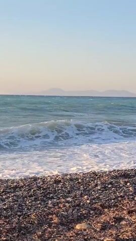 A short video shows waves crashing on a pebble beach. The sky is clear and blue, with a hint of orange at the horizon, suggesting either sunrise or sunset. The water is a turquoise blue, and the waves are white-capped and energetic. The beach is covered in small, smooth pebbles and some dried seaweed. In the background, distant hills or mountains are visible. There are no people or man-made objects in the frame, making it a natural and tranquil scene.