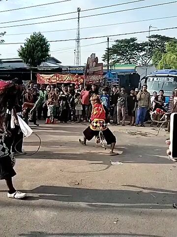 A daytime outdoor cultural performance is taking place on an asphalt road in what appears to be a town or village in Sragen, Indonesia. Several performers, dressed in traditional attire, are engaged in a dynamic dance. Some performers wear elaborate headwear and costumes, including elements that resemble horse figures, suggesting a Jathilan or Kuda Lumping performance. They are holding decorative shields or fans, and one performer wields a whip or rope.

A large crowd of spectators gathers in the background, observing the performance. Buildings, trees, and a tall communications tower are visible behind them. A blue bus with the partially visible text "SELES" is parked in the background. Banners with some visible Indonesian text, including "LUGAS", hang behind the crowd. The weather is sunny with clear skies, casting distinct shadows on the ground. A person in the foreground is holding a smartphone, seemingly recording the event. The atmosphere appears lively and engaging.